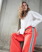 A woman wearing bright coral-red wide-leg track pants with green and white side stripes, paired with a white blouse, posing against a textured concrete wall.