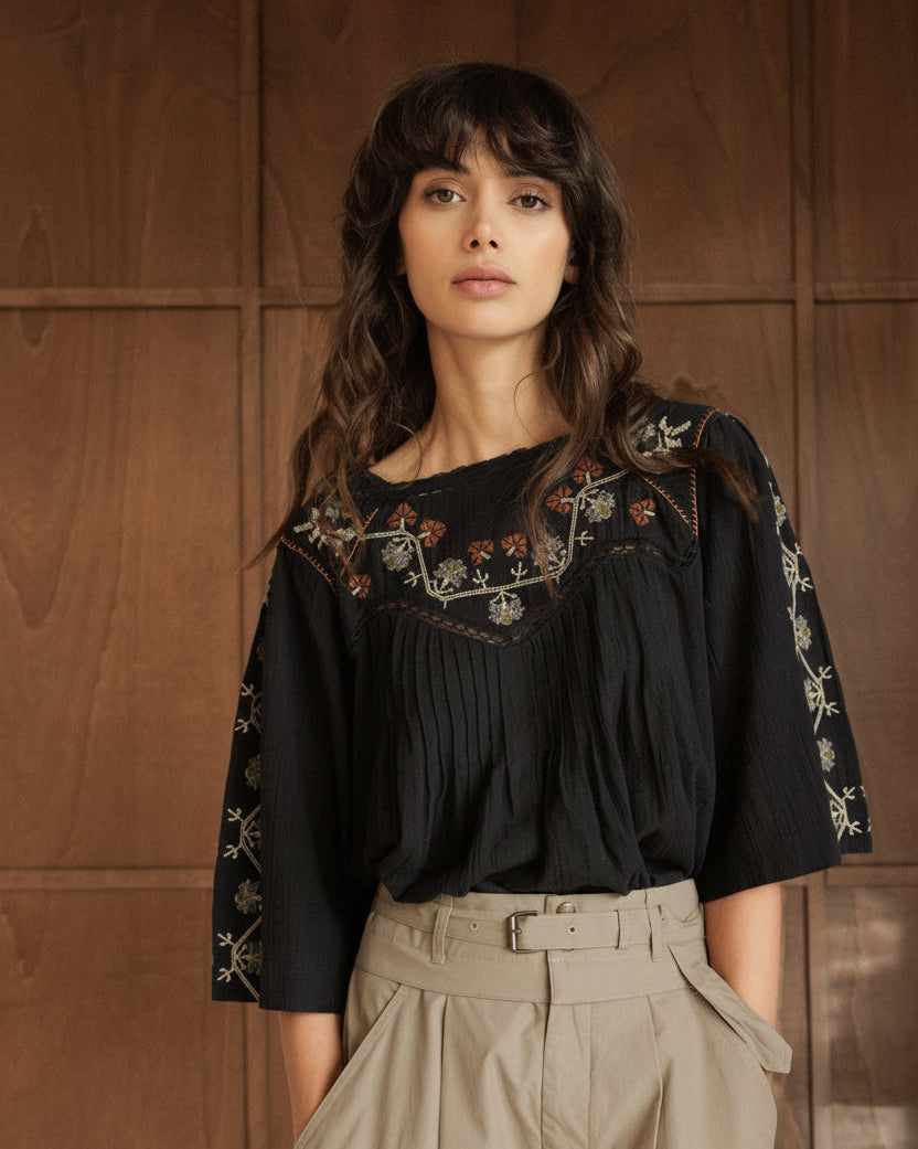 A woman with dark hair and bangs stands against a dark wood-paneled wall, wearing a black embroidered blouse with beige high-waisted trousers.