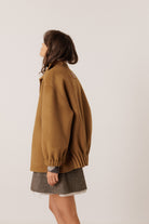A woman shown from the side profile wearing an oversized camel brown jacket and a gray textured skirt, photographed against a plain light gray studio background.