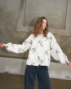 A woman modeling a white long-sleeve blouse with a delicate floral pattern and dark wide-leg jeans, standing against a textured concrete wall.