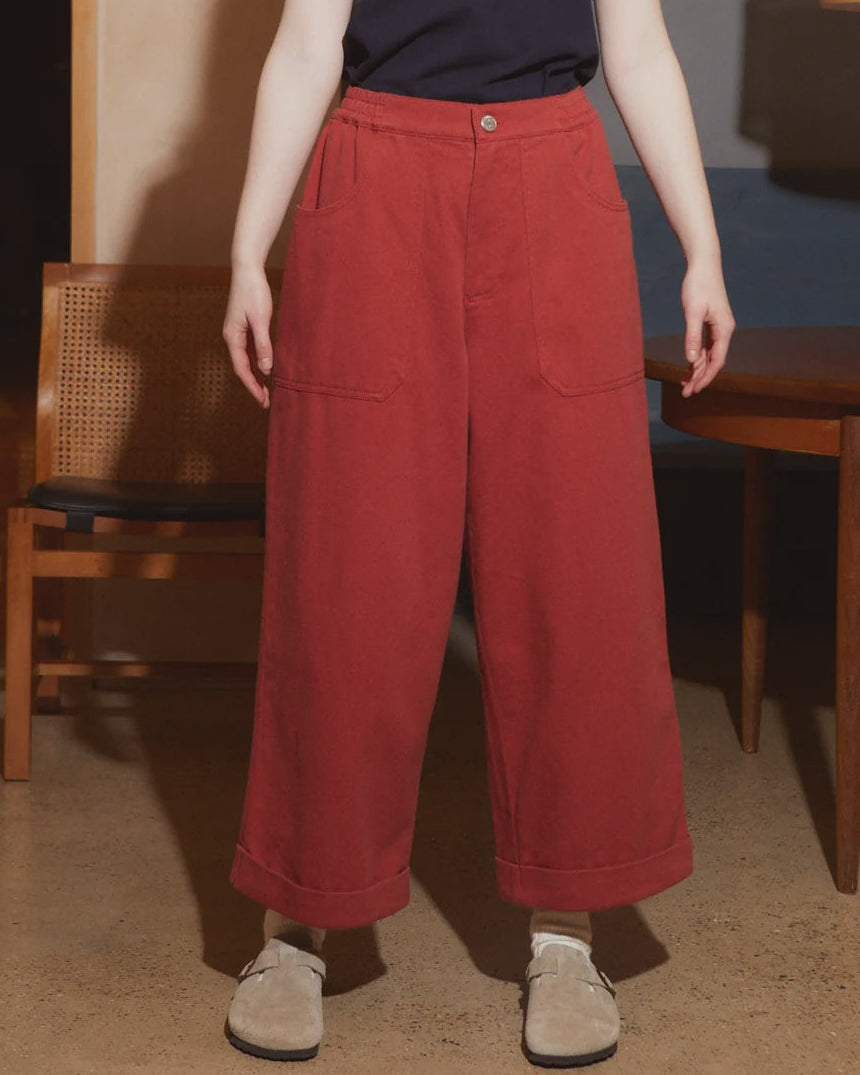 A model wearing a dark navy t-shirt and high-waisted, wide-leg red corduroy pants, standing in a modern indoor setting with wooden furniture and a hanging lamp.