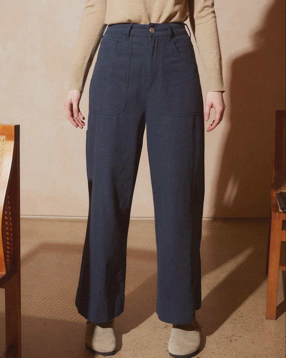 A model wearing high-waisted navy blue wide-leg trousers, paired with a beige long-sleeve top and beige slippers, standing in a warm-toned room.
