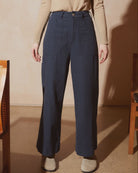A model wearing high-waisted navy blue wide-leg trousers, paired with a beige long-sleeve top and beige slippers, standing in a warm-toned room.