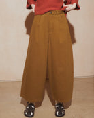 A person wearing a rust-red ribbed knit sweater and wide-leg mustard-brown trousers, standing indoors against a plain beige wall with sandals visible at the bottom.