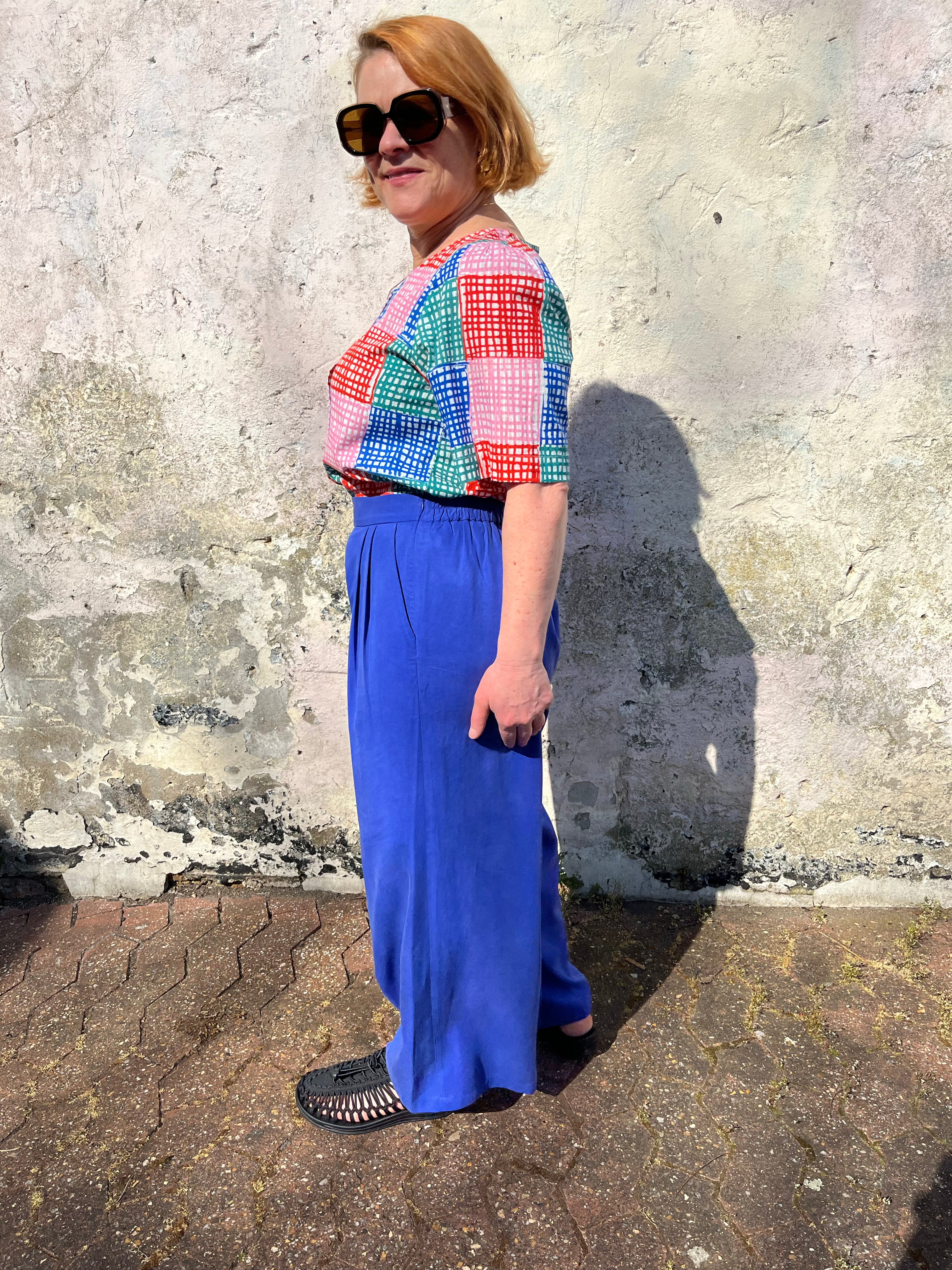 A person standing against a wall wearing cobalt lyocell wide-leg trousers with a flat front waistband, elasticated back, and pleats at the front.