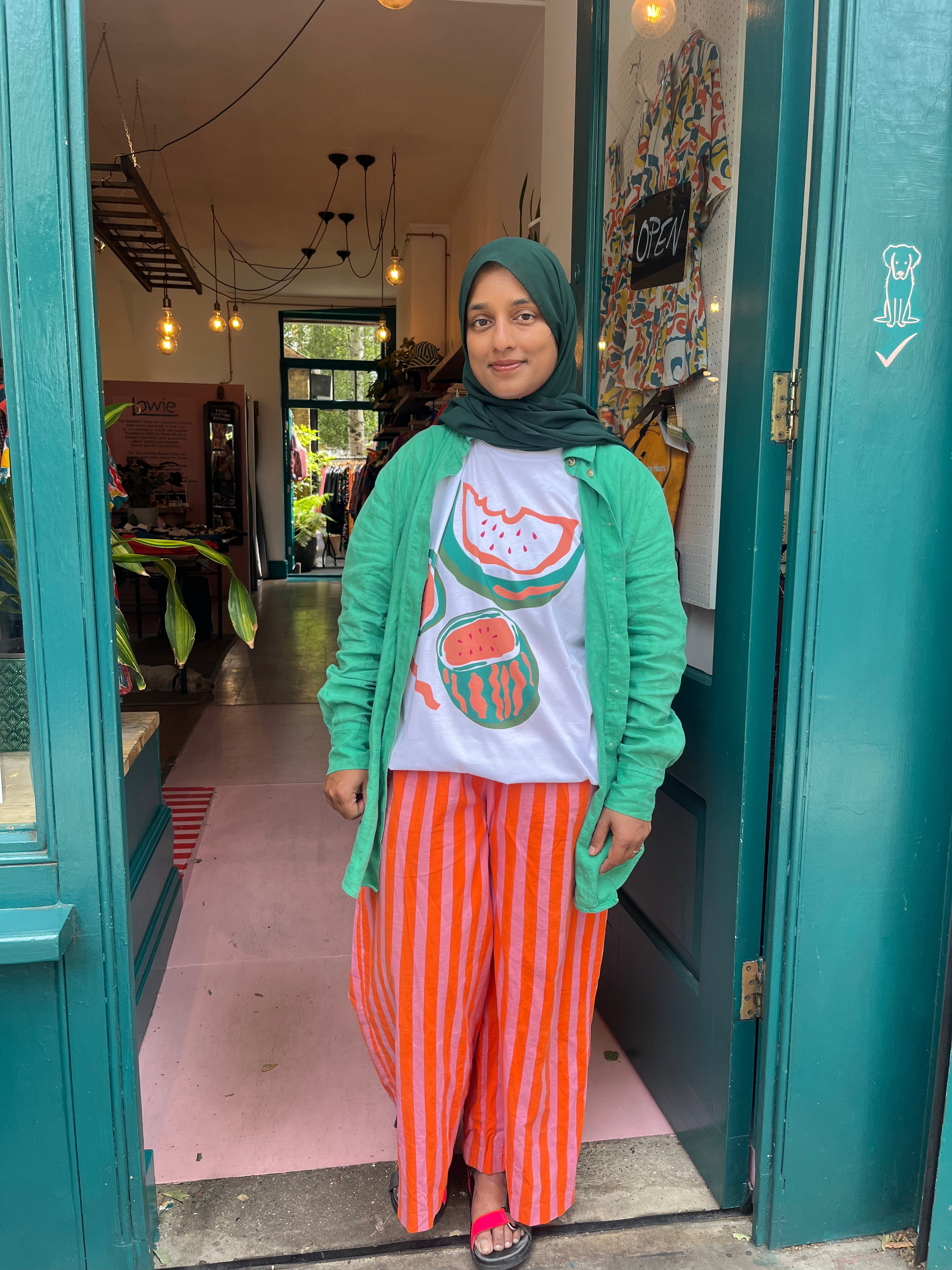 Woman standing in a doorway wearing a green cardigan, white shirt with graphic design, and orange striped pants.
