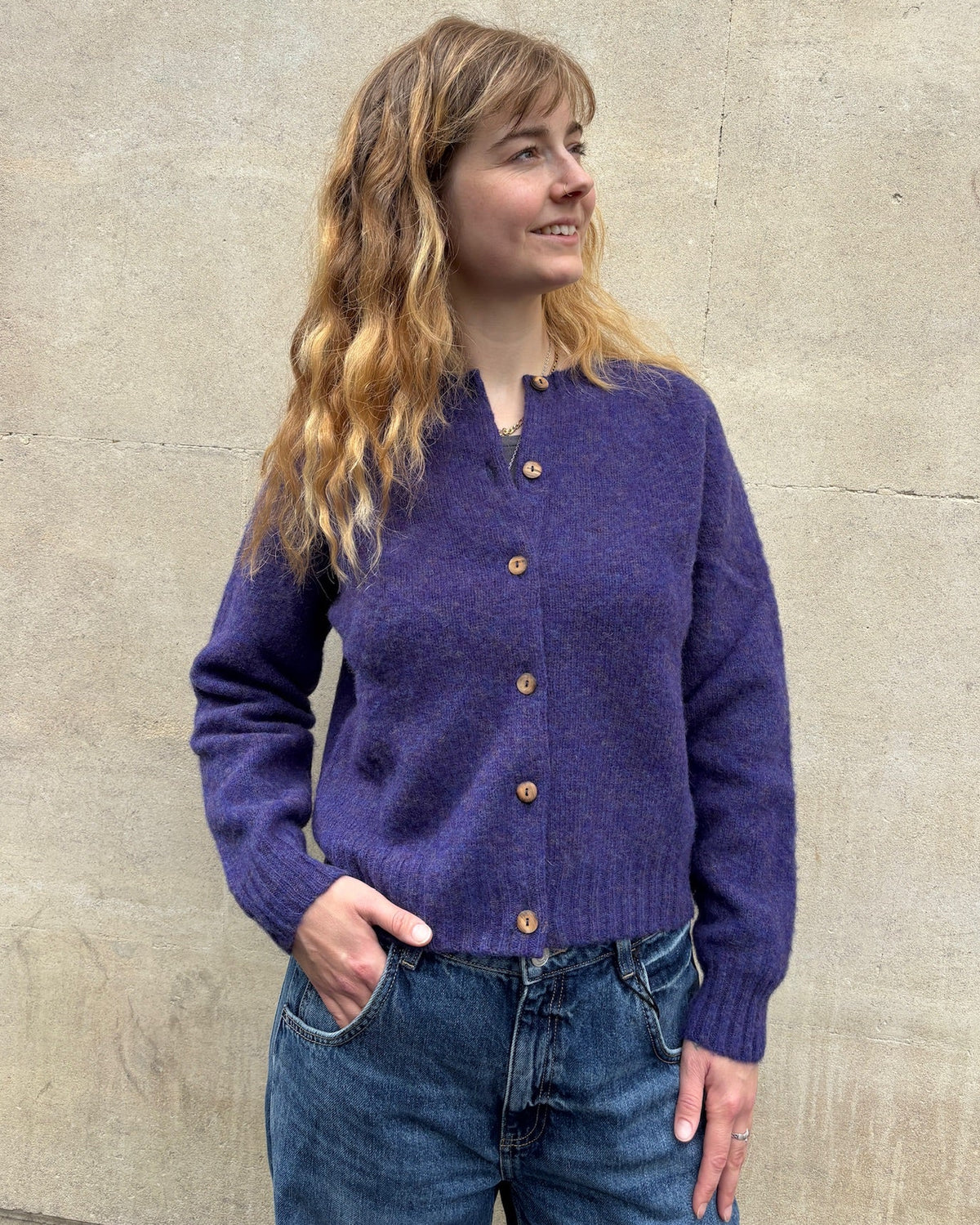 A woman with long wavy light brown hair wearing a purple knit button-up cardigan and blue jeans, standing against a textured concrete wall.