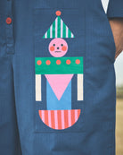 A close-up of a person wearing a blue short-sleeve button-front garment with a large colorful patchwork pocket featuring a stylized face and geometric shapes in green, pink, blue, white, and orange.
