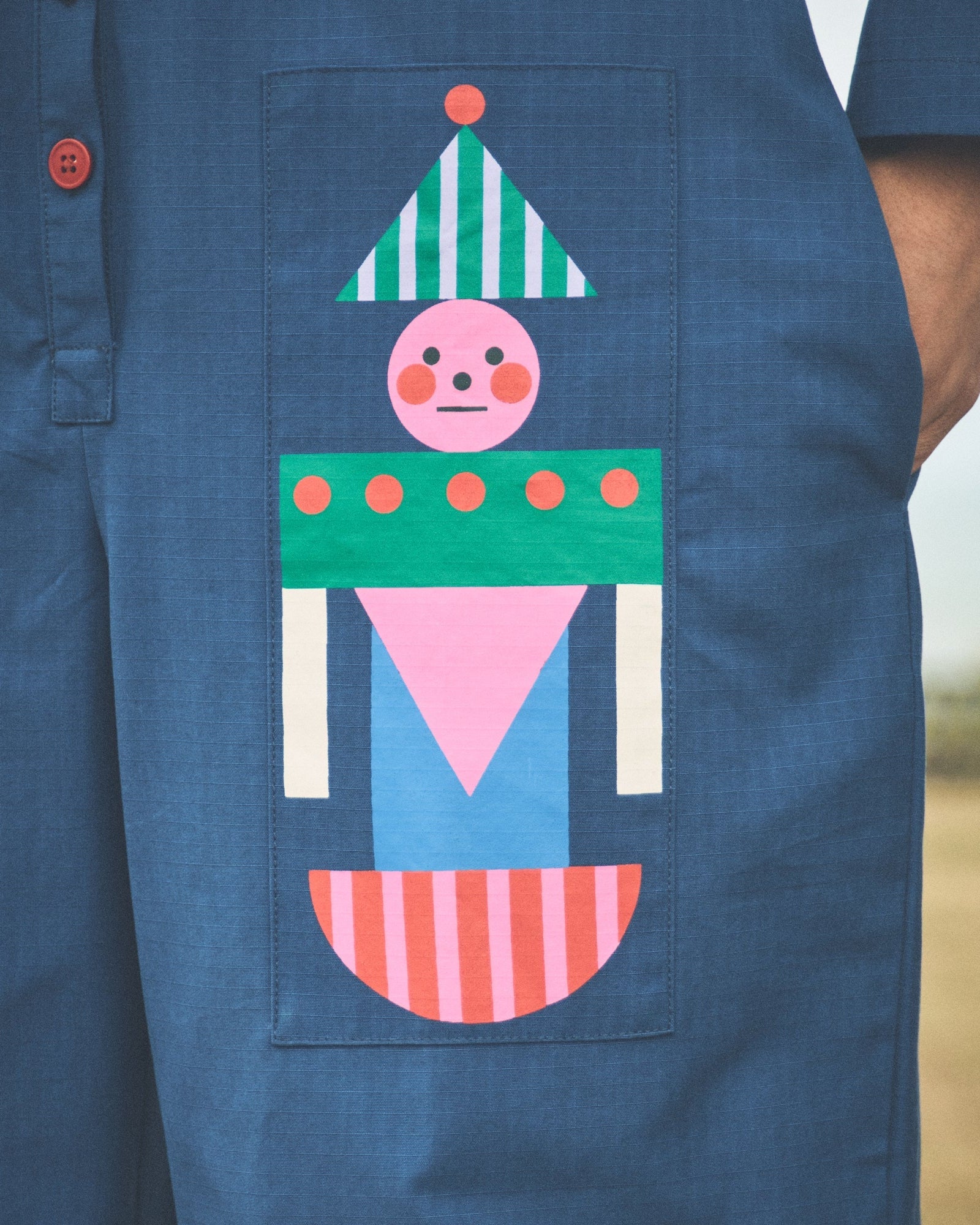 A close-up of a person wearing a blue short-sleeve button-front garment with a large colorful patchwork pocket featuring a stylized face and geometric shapes in green, pink, blue, white, and orange.