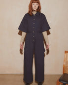 A model with red hair stands against a beige wall wearing a dark navy wide-leg jumpsuit with short sleeves and a button-up front, layered over a beige long-sleeve shirt.