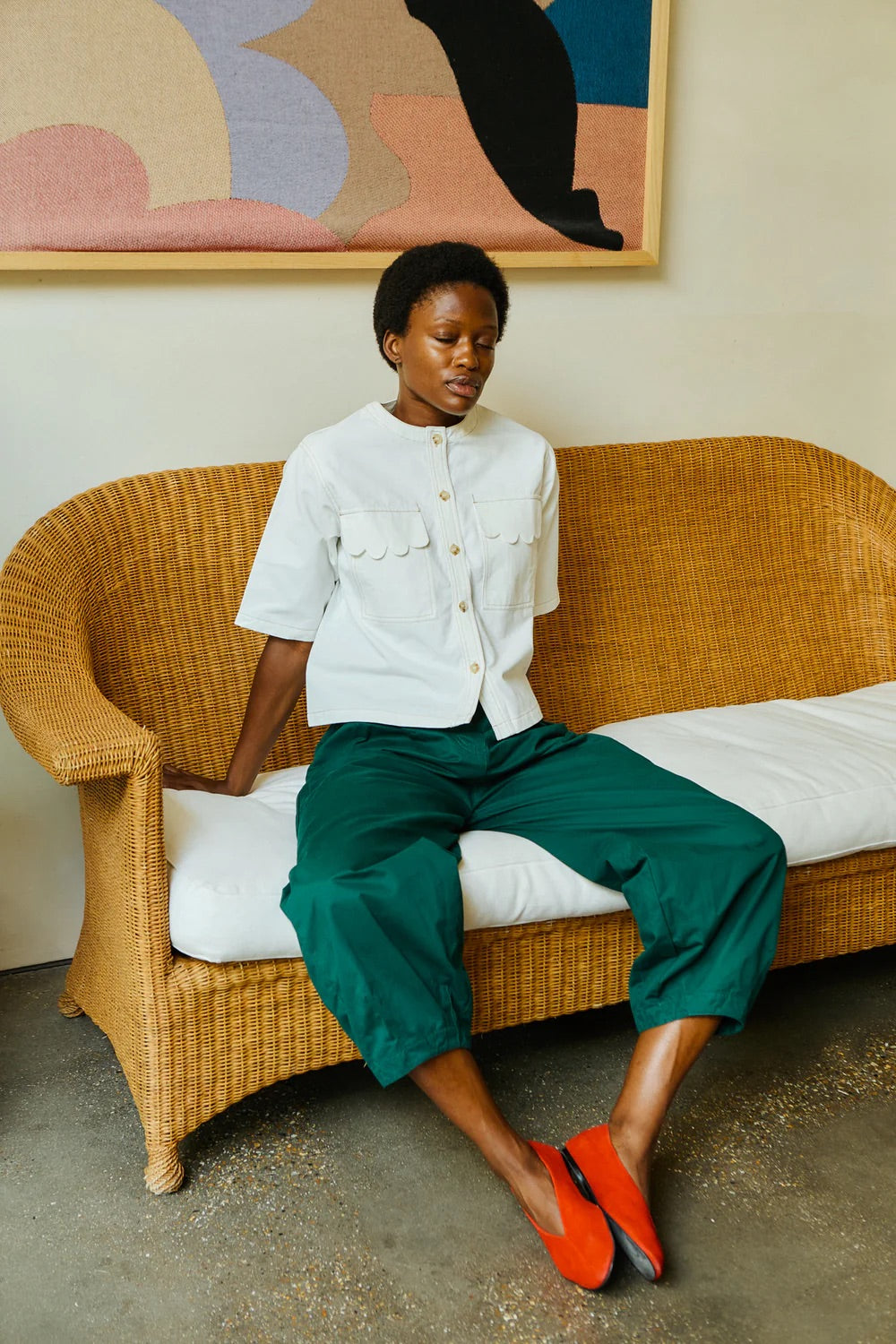 Woman sitting on a wicker couch wearing a white jacket and green pants, with a colorful abstract painting in the background.
