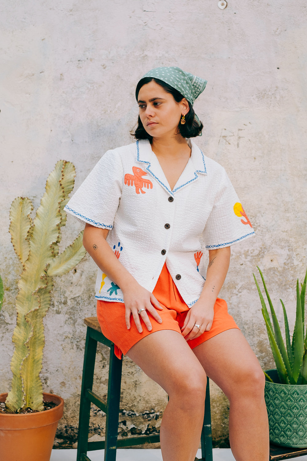 A white cotton blouse with colorful hand embroidery details, featuring a notched collar, wide cut sleeves, and a boxy fit.