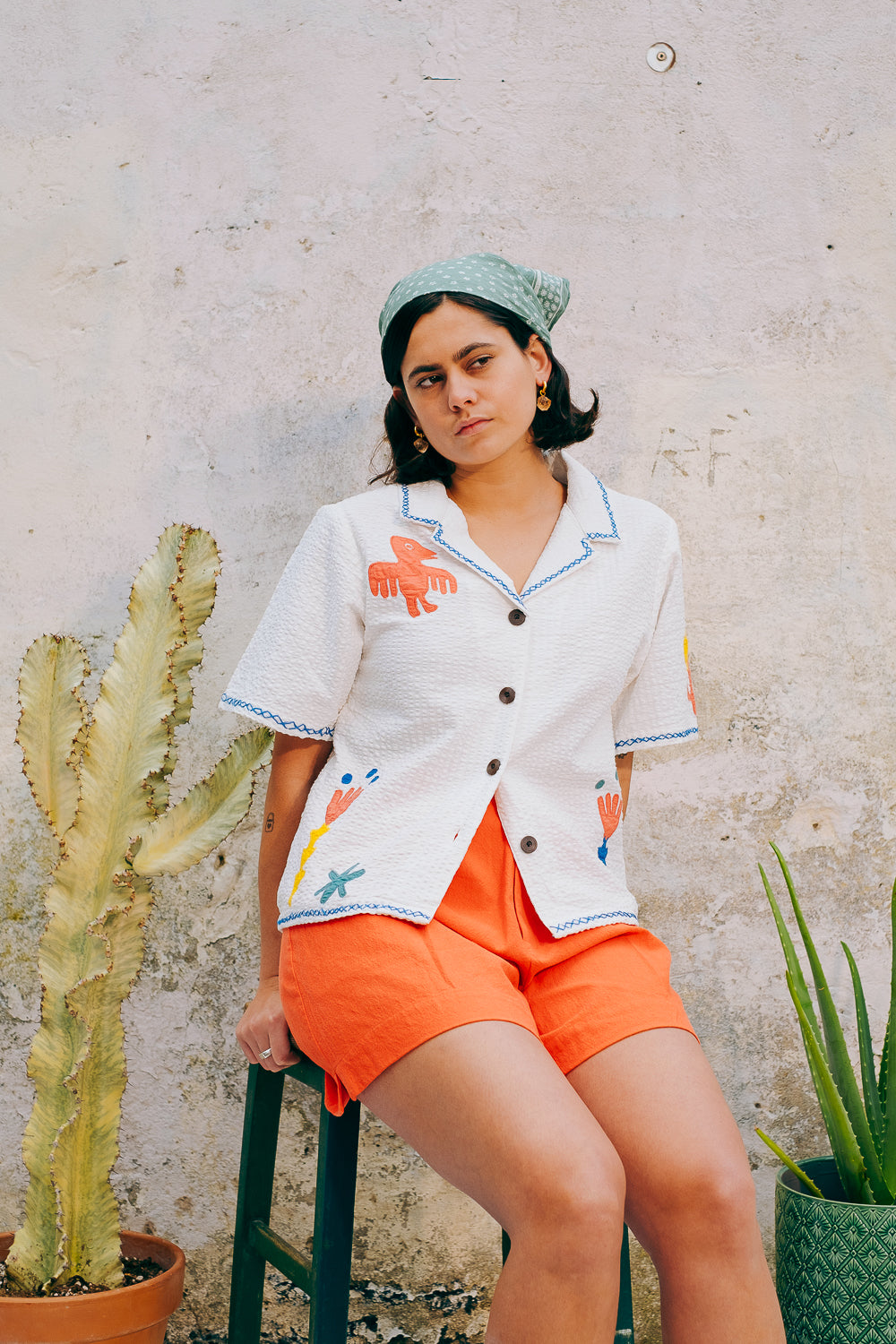 A white cotton blouse with colorful hand embroidery details, featuring a notched collar, wide cut sleeves, and a boxy fit.