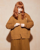 A model with long red hair wearing a brown corduroy jacket and matching wide-leg trousers, standing against a beige textured wall.