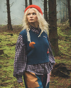 A model stands in a misty, mossy forest wearing a blue and red patchwork sweater vest over a blue-and-white gingham shirt, paired with blue pants featuring a tan patch and a red beret.