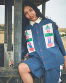 A woman wearing a blue denim-like short-sleeve button-front top and matching shorts with colorful patchwork applique patches, posed outdoors against a wooden structure.