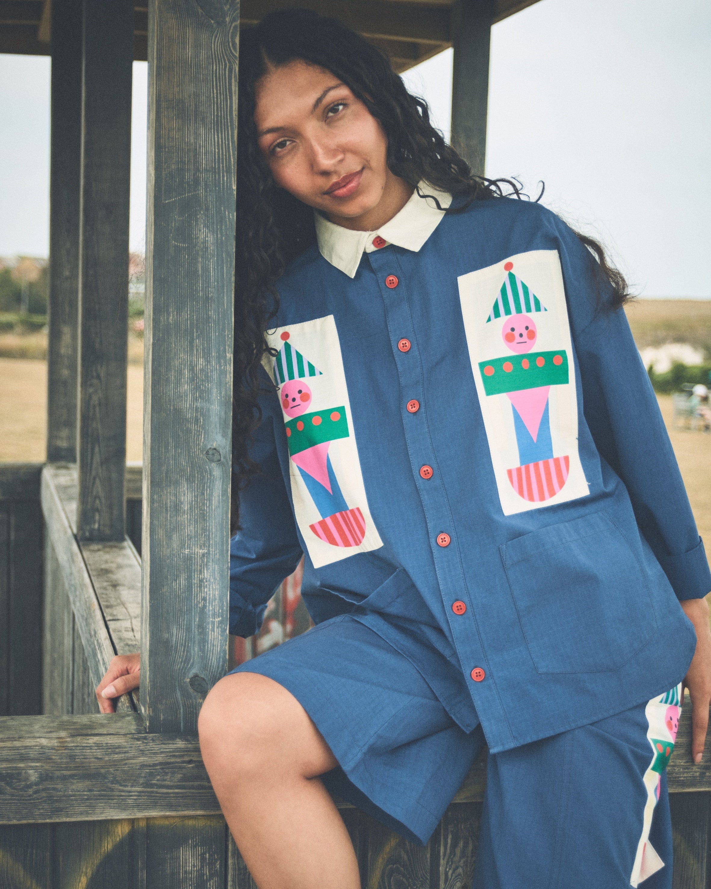 A woman wearing a blue denim-like short-sleeve button-front top and matching shorts with colorful patchwork applique patches, posed outdoors against a wooden structure.