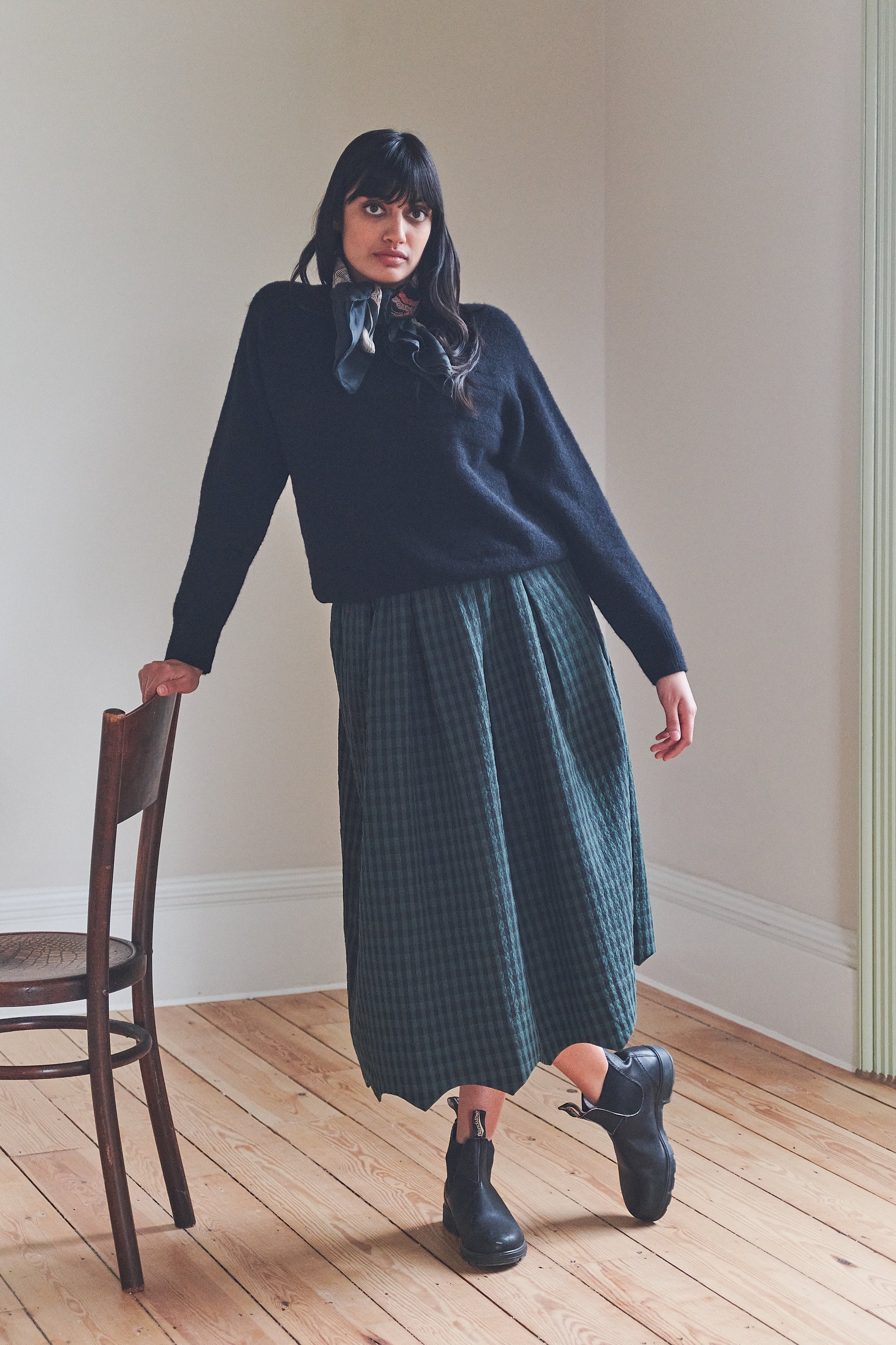 Woman wearing a navy sweater and green plaid skirt standing in a room with wooden floor and white walls.