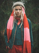 A woman wearing a chunky knit beanie, a teal button-front cardigan, a bright pink and orange striped scarf, and rust-red pants, standing outdoors against a dark forest background.
