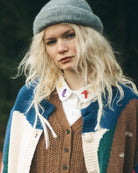 A woman stands in a forest wearing a chunky knit color-block cardigan, brown knit vest, white shirt, dark trousers, and a gray beanie.