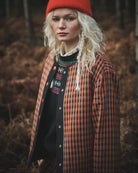 A model wearing a bright red knit beanie, a rust-orange and black checkered overshirt, and a black knit sweater with colorful embroidery, standing in a forest with dry brown foliage.