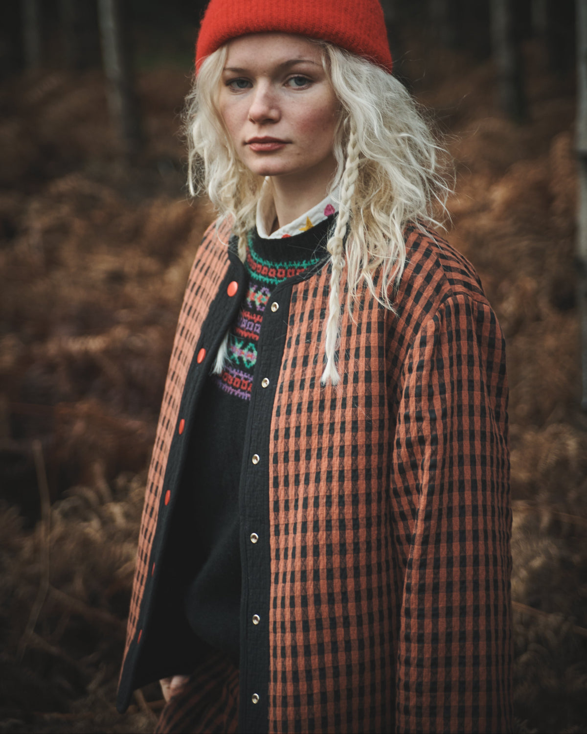 A model wearing a bright red knit beanie, a rust-orange and black checkered overshirt, and a black knit sweater with colorful embroidery, standing in a forest with dry brown foliage.