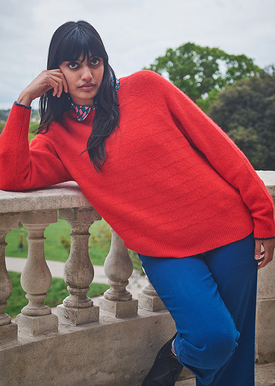 Person wearing a bright red sweater and blue pants standing on a stone railing with greenery in the background