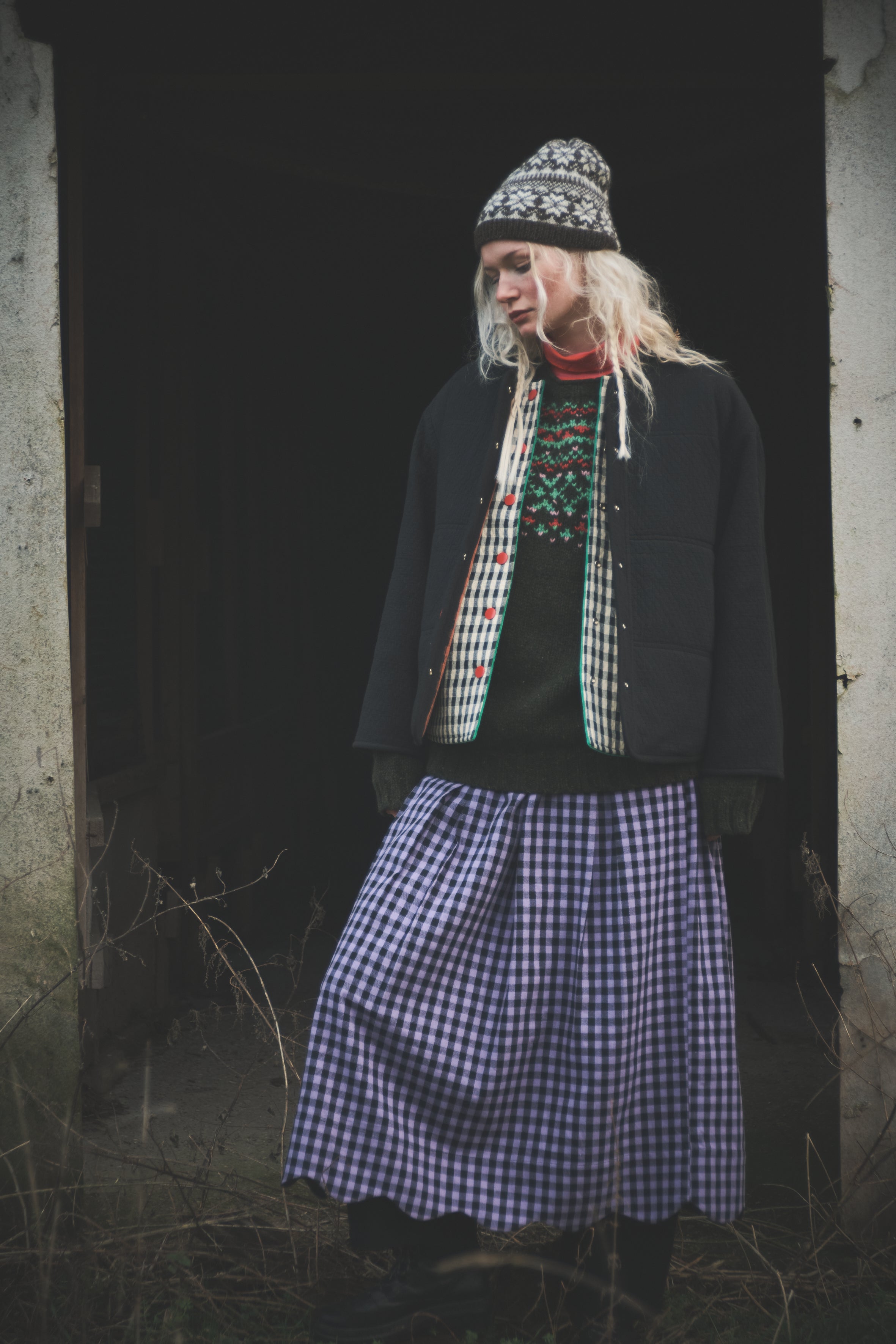 A person standing in a dark doorway wearing a black jacket, a patterned knit beanie, a red and green patterned sweater, and a blue-and-white gingham skirt.