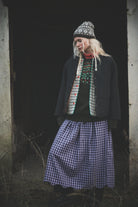 A person standing in a dark doorway wearing a black jacket, a patterned knit beanie, a red and green patterned sweater, and a blue-and-white gingham skirt.