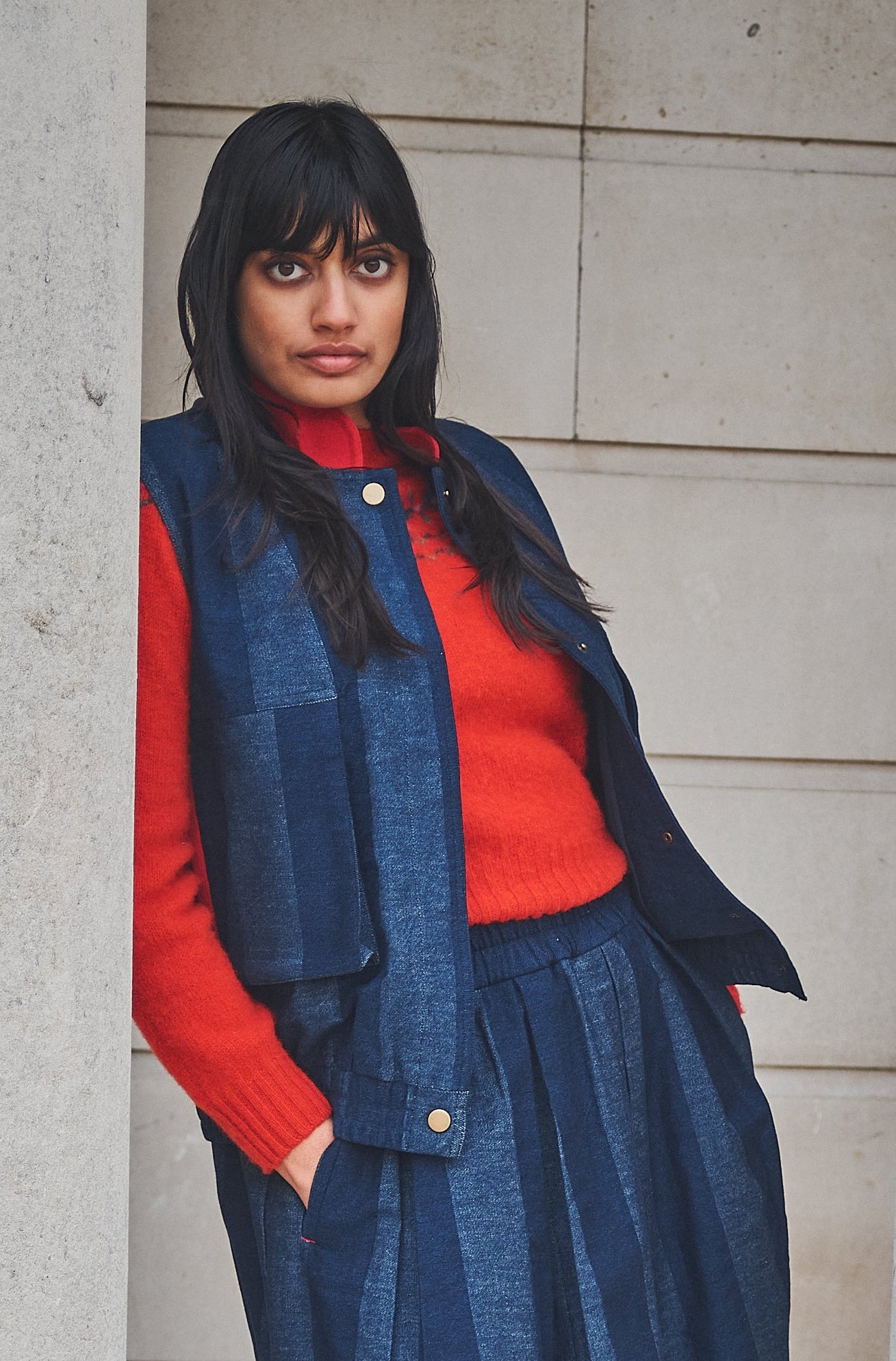 Woman in a denim gilet, red fair isle jumper and blue skirt standing between stone columns.