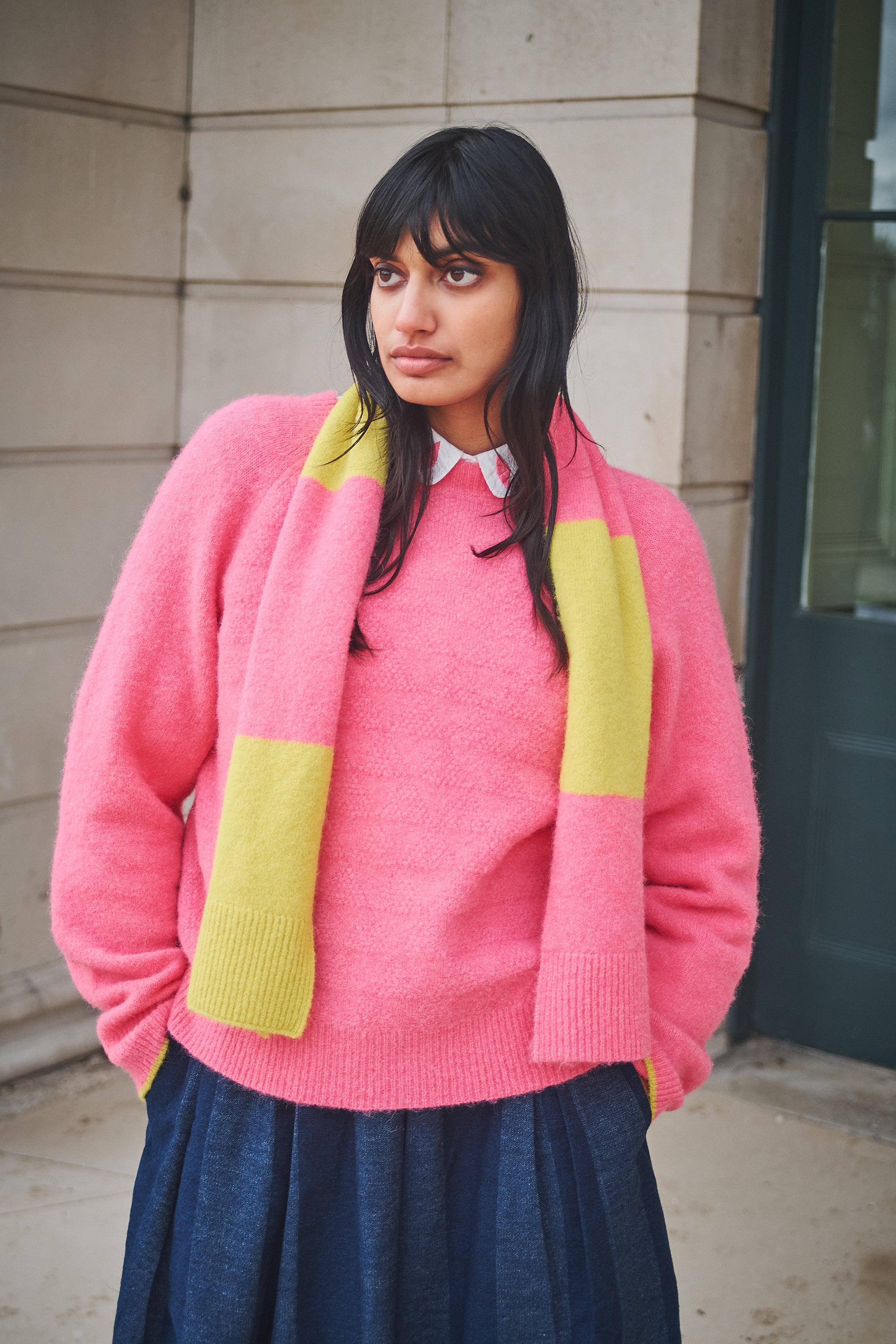 Woman wearing a pink sweater with yellow accents standing against a building.