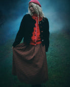 A woman stands outdoors in a misty blue-green environment wearing a bright pink beret, a black cardigan over a vibrant orange-red sweater, and a brown and black checkered skirt.