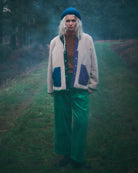 A person standing on a grassy path in a misty forest, wearing a cream cardigan with blue patch pockets, bright green pants, and a blue beret.