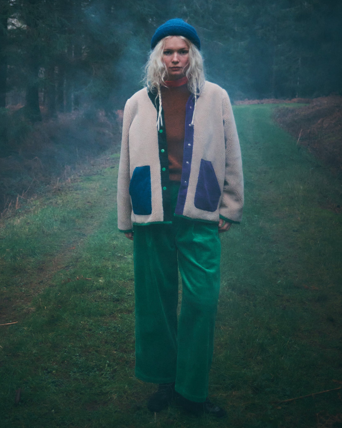 A person standing on a grassy path in a misty forest, wearing a cream cardigan with blue patch pockets, bright green pants, and a blue beret.