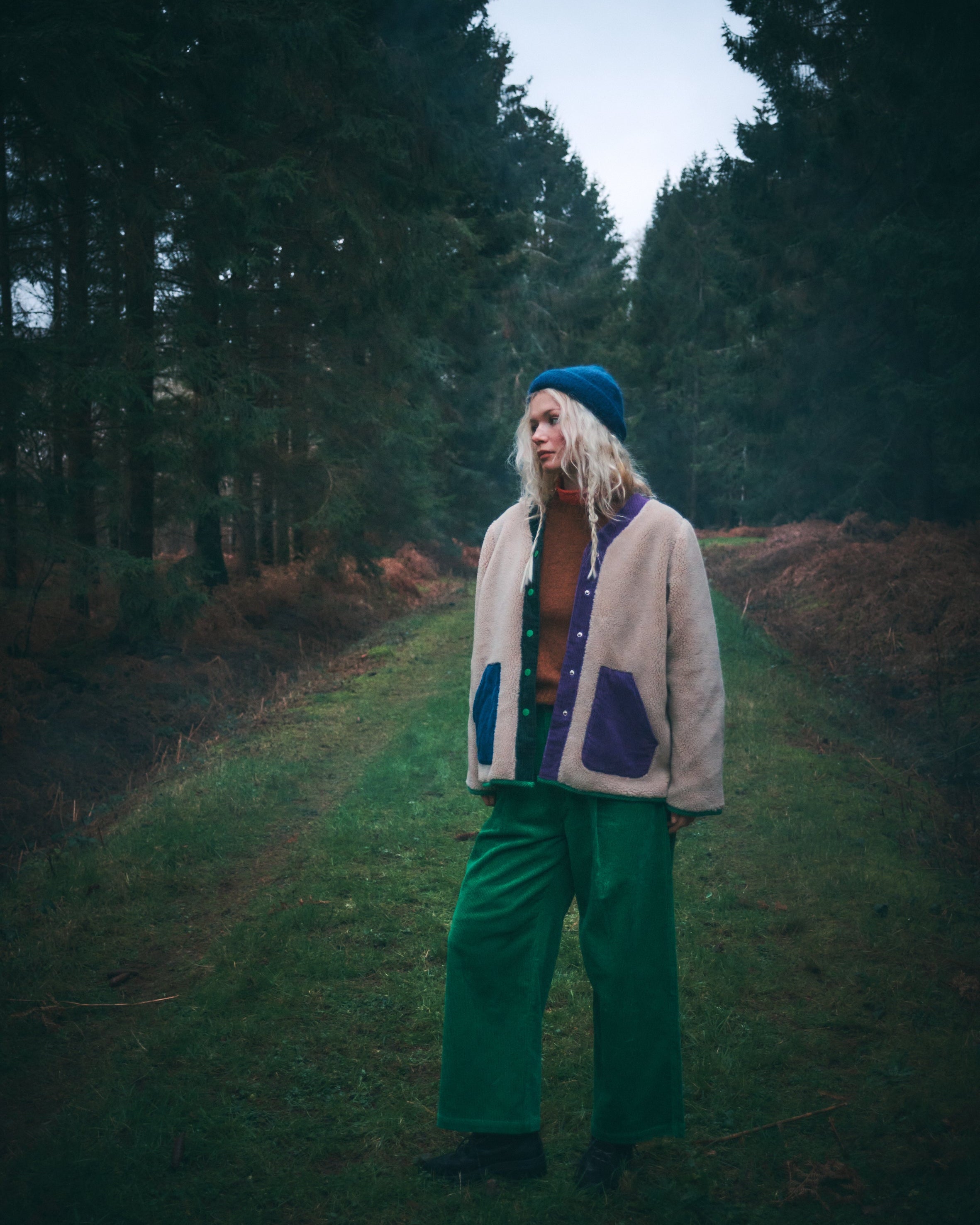 A person standing on a grassy path between dark pine trees, wearing a beige and purple fleece jacket, green wide-leg pants, a brown top, and a blue beanie.