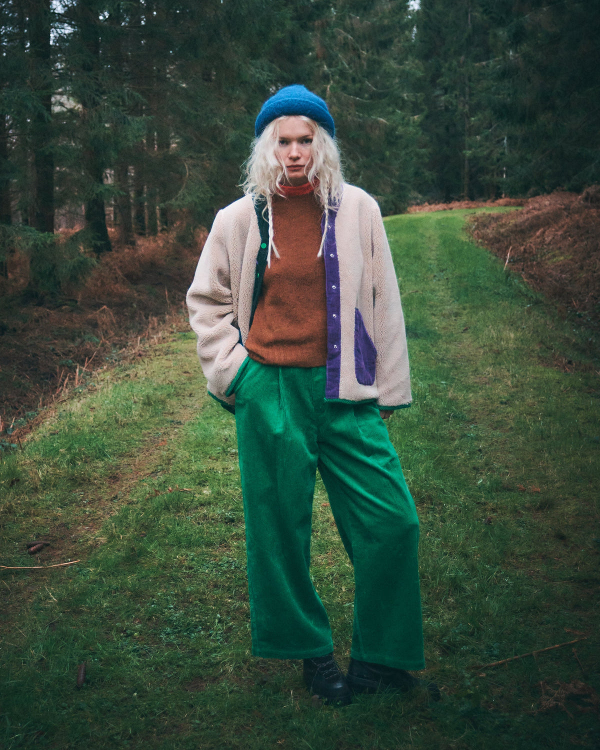A person standing on a grassy path in a forest, wearing a blue beret, a cream cardigan with purple and orange panels, a rust-orange turtleneck, and bright green wide-leg pants.