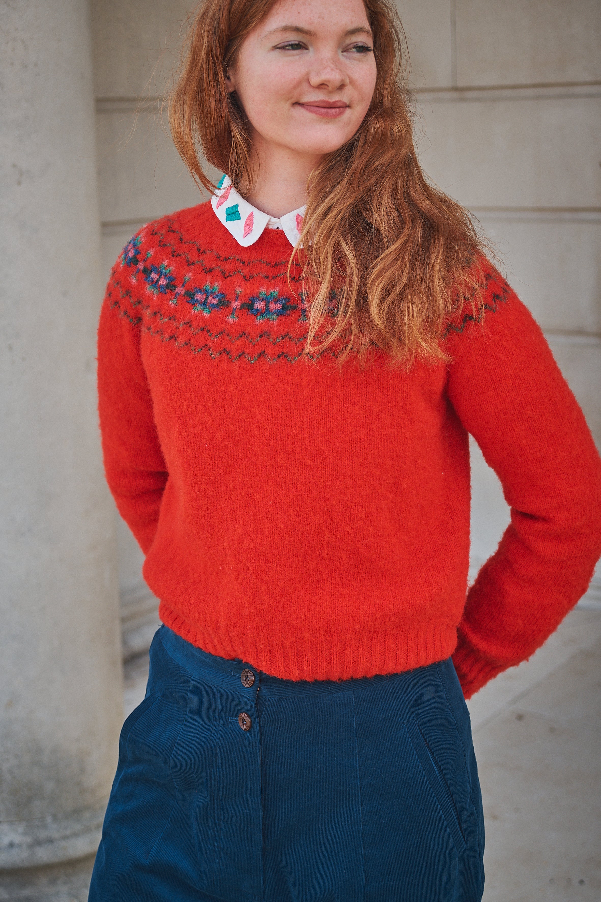 Woman wearing a red knitted sweater with floral patterns against a stone wall.