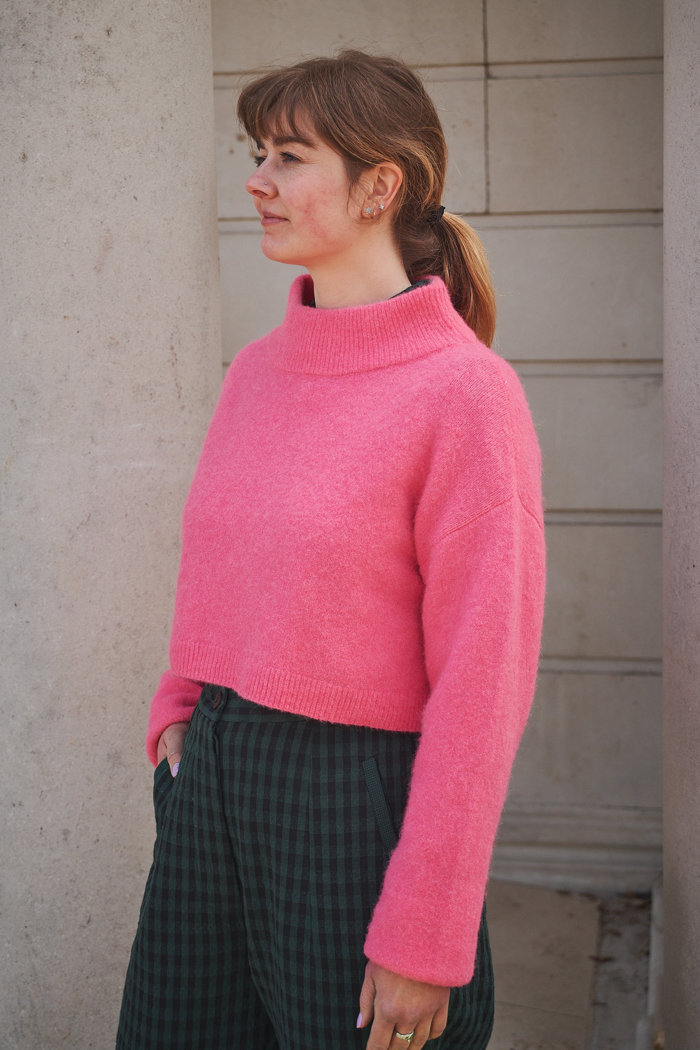 Person wearing a pink sweater and plaid pants standing against a neutral wall.