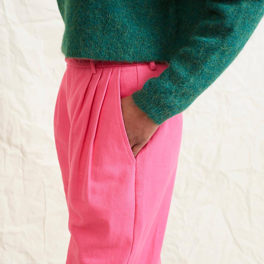A model wearing bright pink wide leg trousers with pleats and a tab front opening, paired with a green sweater and brown boots.