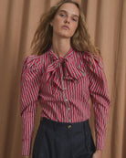 A woman wearing a red and white vertically striped long-sleeve blouse with ruffled details and a bow at the collar, paired with dark navy trousers, standing in front of a draped tan curtain.