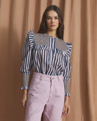 A model stands in front of a draped tan fabric backdrop wearing a navy and white vertically striped blouse with ruffled shoulders and light pink wide-leg jeans.