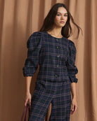 A woman models a matching navy blue plaid two-piece set with puff sleeves and wide-leg trousers, styled with a dark purple handbag and metallic flats, set against a draped tan fabric backdrop.