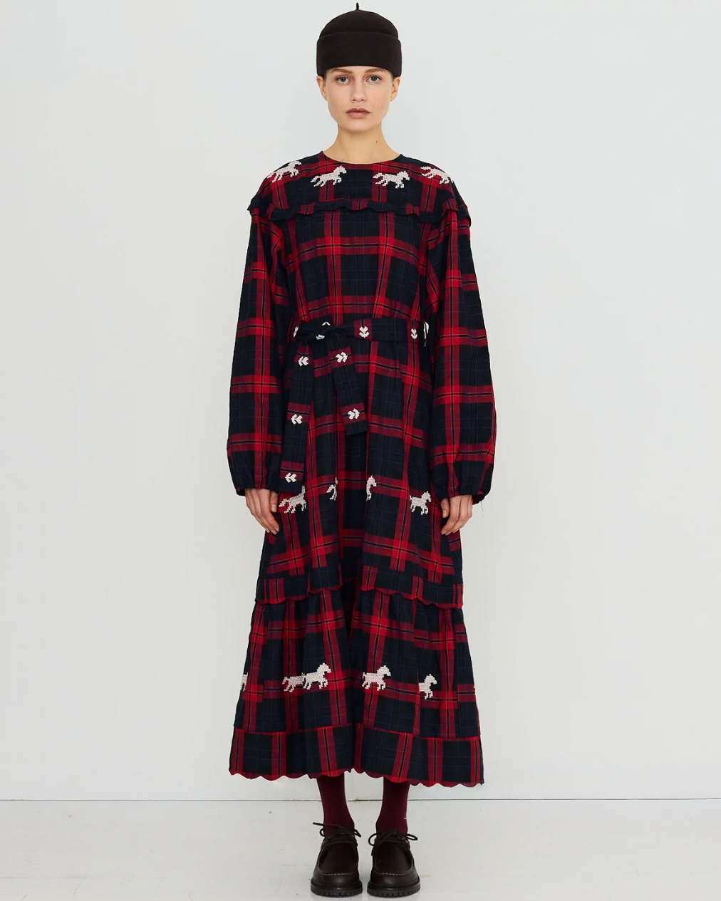 A model stands against a plain white studio background wearing a long-sleeved, knee-length plaid dress with a red and black tartan pattern and scattered white dog silhouettes. The dress has a matching tie belt at the waist and a ruffled hem.