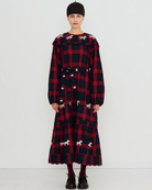 A model stands against a plain white studio background wearing a long-sleeved, knee-length plaid dress with a red and black tartan pattern and scattered white dog silhouettes. The dress has a matching tie belt at the waist and a ruffled hem.