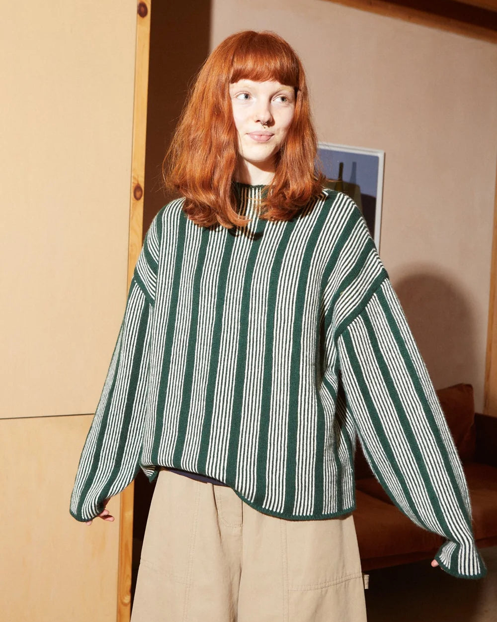A woman with red hair wearing a green and white vertically striped cropped sweater and beige wide-leg pants, standing in a warm-toned indoor room.
