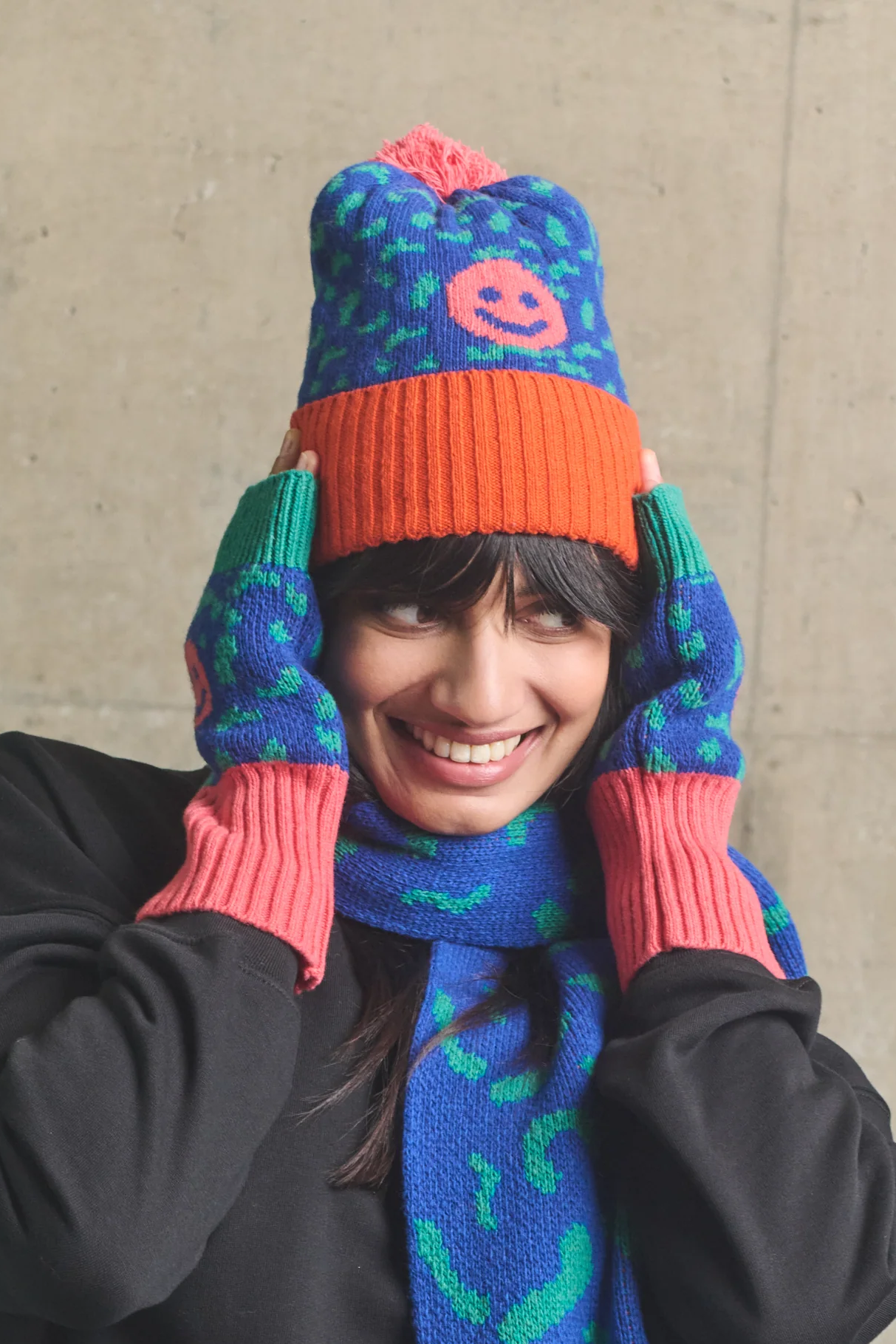 Person wearing a colorful knitted hat and scarf against a concrete wall.