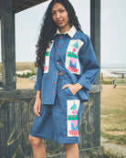 A woman stands outdoors in front of a wooden structure at a grassy coastal location, wearing a blue matching shirt and skirt set with large multicolor geometric patchwork panels on the front.