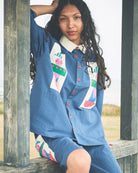 A woman wearing a blue matching lounge set with colorful geometric and cartoon-style patchwork prints, sitting on a wooden structure outdoors.
