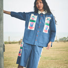 A woman wearing a blue denim-like matching set with colorful geometric patchwork details on the chest and sleeve, photographed outdoors near a grassy field and water.