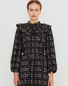 A model wearing a long-sleeved black dress with a beige floral grid pattern, featuring a high ruffled collar and a matching headband, set against a plain light gray background.